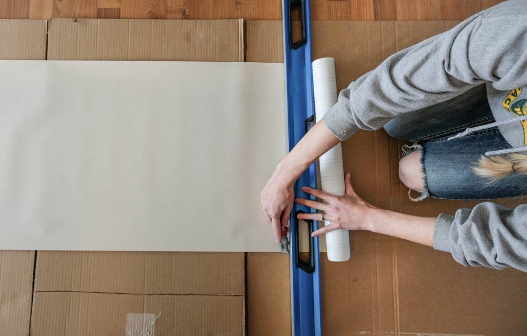 Overhead view of someone using a level and boxcutter to cut a strip of beadboard wallpaper