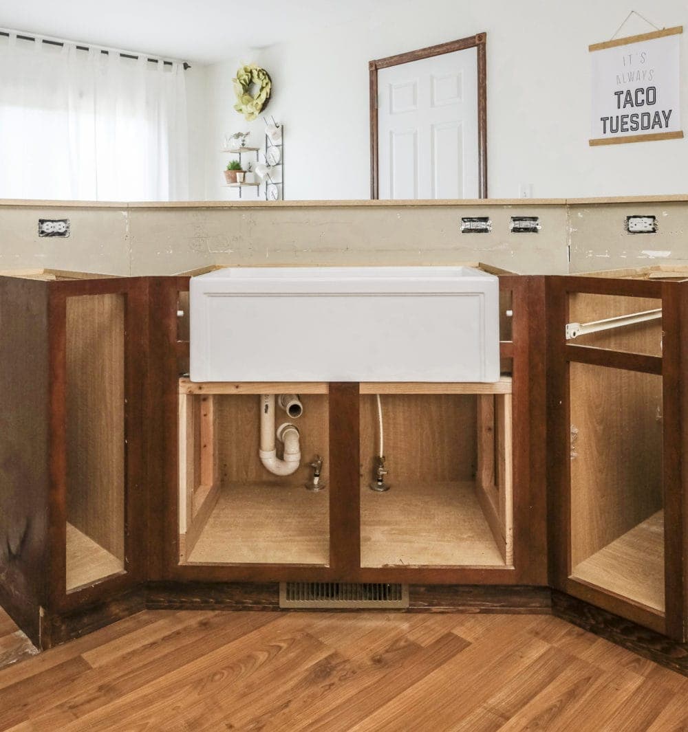 View of cabinets with doors removed and a white apron front farmhouse sink sitting in place before counterops installed