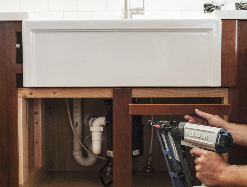 Nail gun installing cabinet trim piece under farmhouse sink