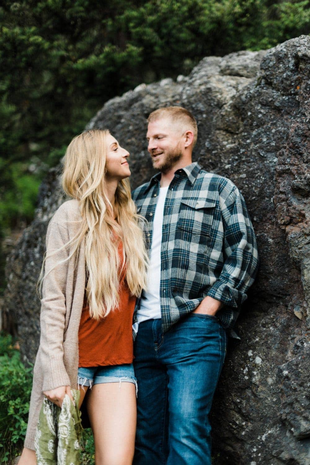 Blonde couple looking at each other leaning on a large rock in the woods embracing RV life