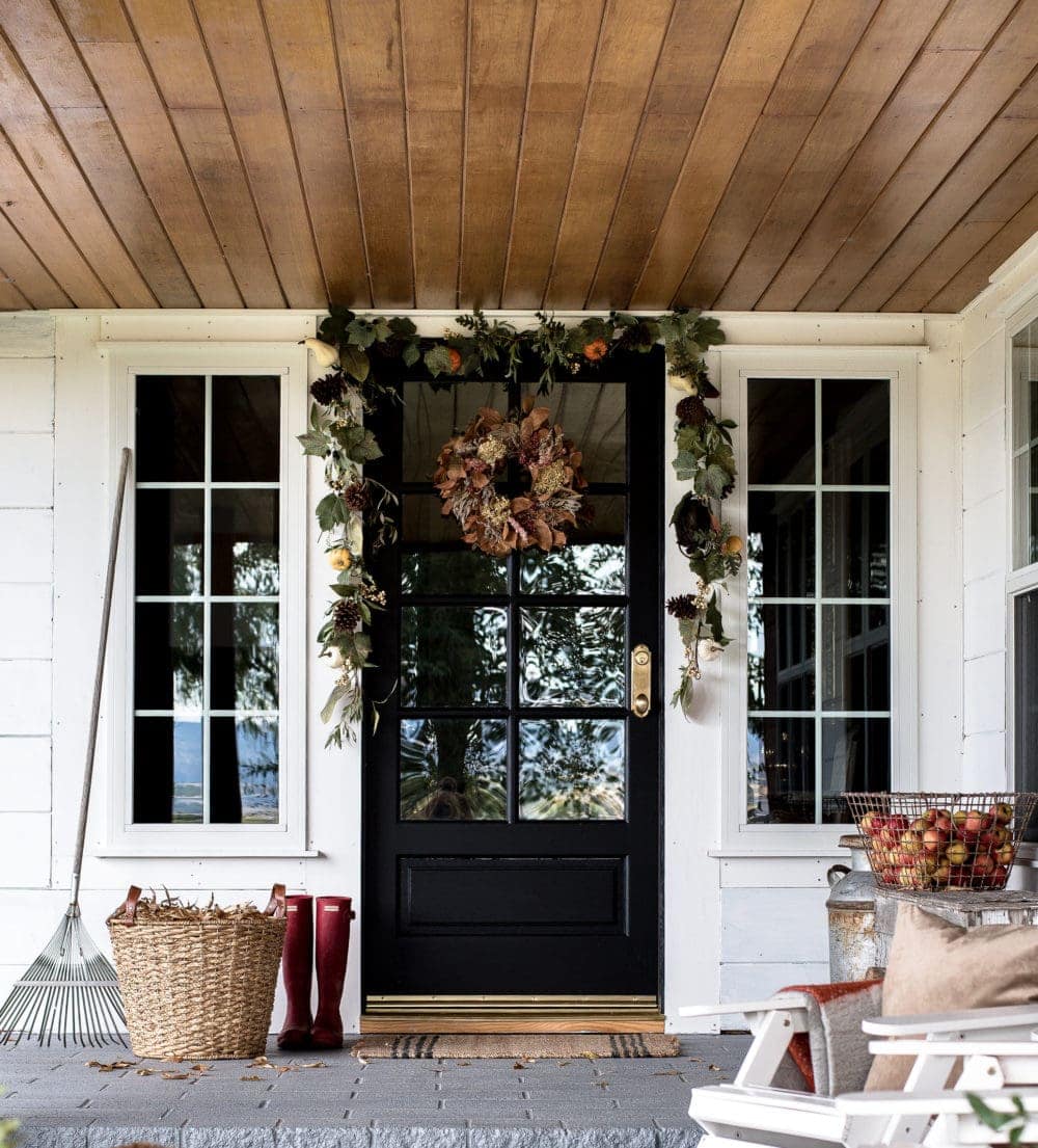 Fall front porch ideas using a basket of dried leaves next to a rake, a basket of apples, and a green garland over the door