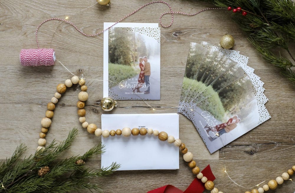 Personalized Christmas cards layed out next to envelopes surrounded by ribbon, garland and greenery