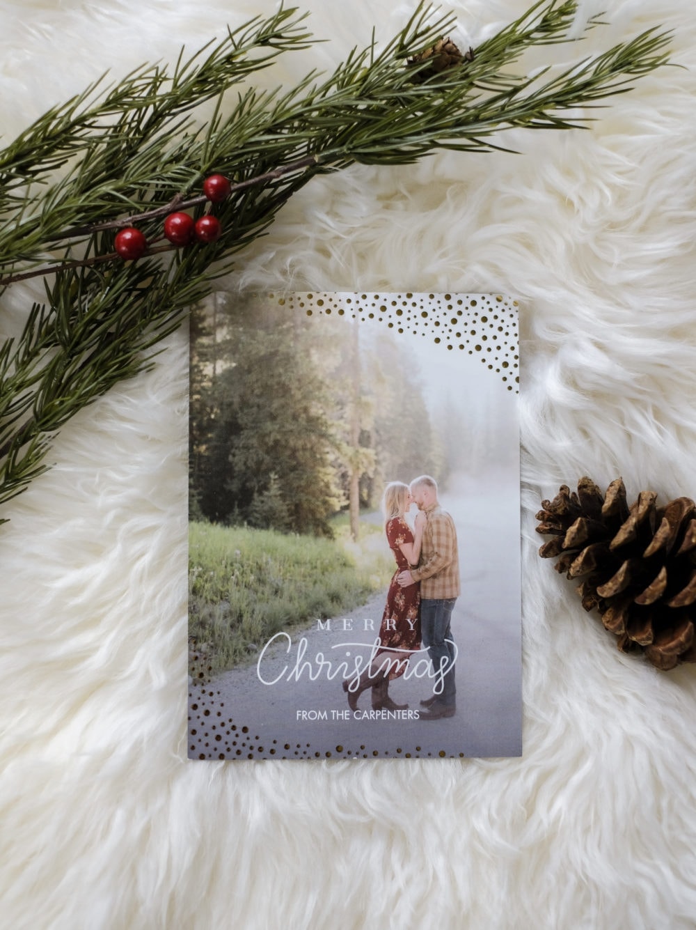 A personalized Christmas card laying on a white sheepskin rug next to greenery and a pinecone