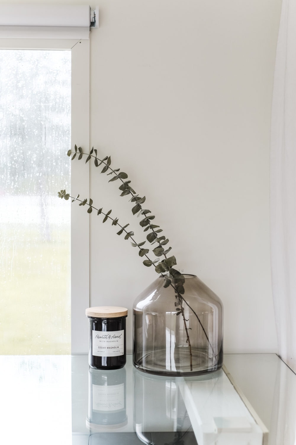 A glass vase with dried eucalyptus next to a candle on top of a glass desktop